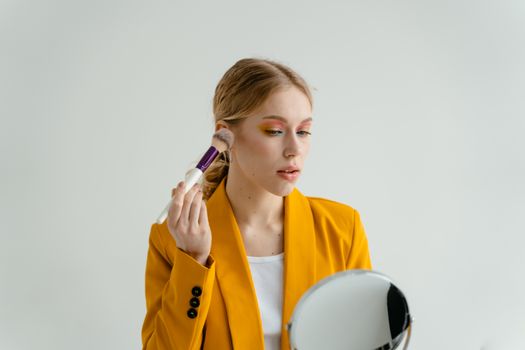 Stylish woman in a yellow blazer applying makeup using a mirror indoors.