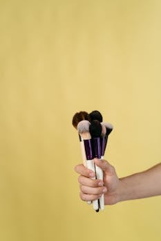Closeup of hand holding assorted makeup brushes with pastel yellow backdrop.