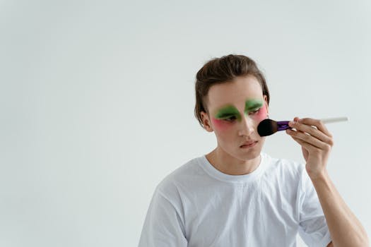 Young man in a white shirt applies vibrant makeup using a brush against a plain background.