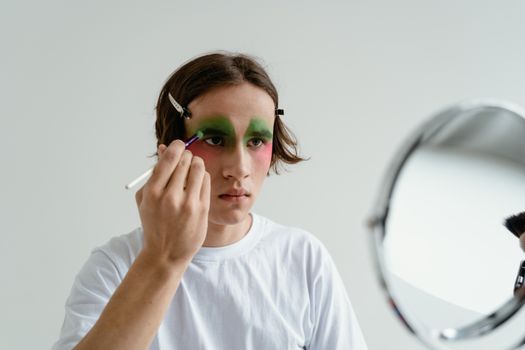 A young man applies bold green and red makeup while looking into a mirror, showcasing creativity.