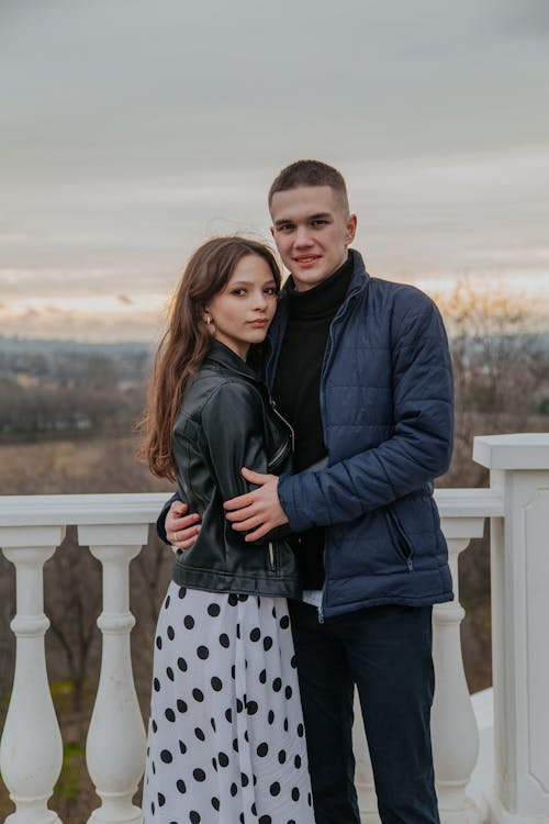 Free Loving young couple in outerwear looking at camera while embracing on street near while balustrade during romantic date against grassy field Stock Photo