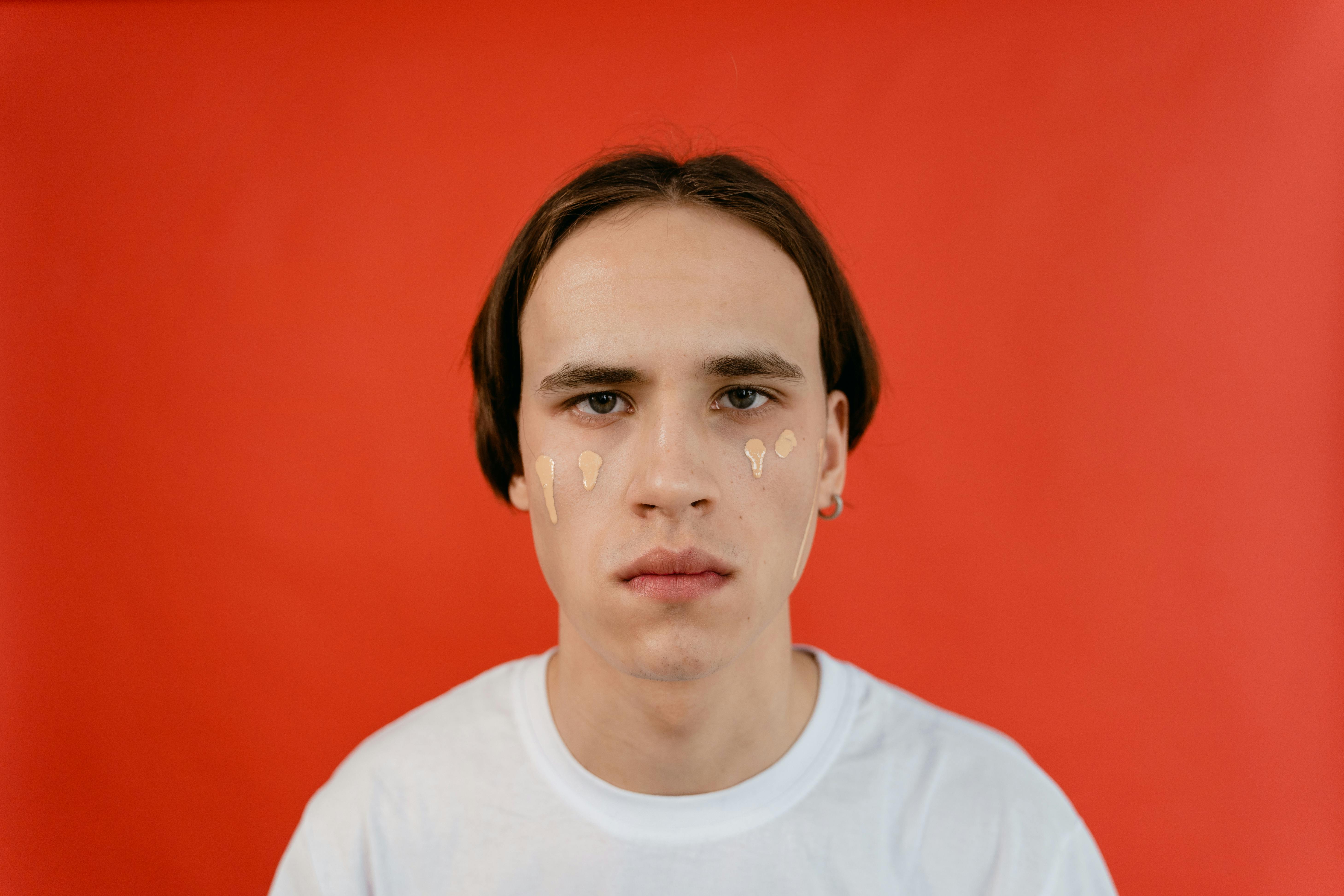 A young man applying liquid foundation on his face in a studio with red background.