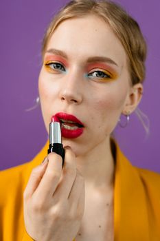Close-up of a woman applying red lipstick with colorful eye makeup against a purple background.