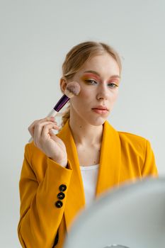 Caucasian woman in yellow blazer applying makeup with brush in front of mirror.