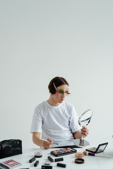 A man sitting indoors, applying makeup using a brush and mirror, surrounded by cosmetics.