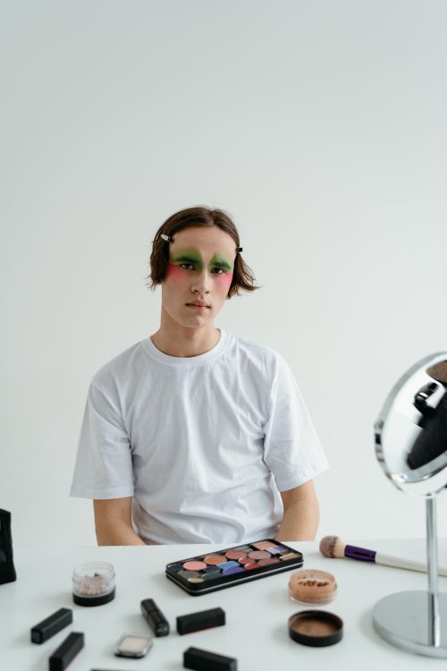 Free Young man with vibrant makeup sits by a table of cosmetics, embodying artistic expression. Stock Photo