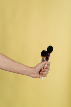 A close-up of a hand holding makeup brushes against a yellow backdrop.