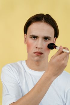 A young man holding a makeup brush to his face against a vibrant yellow background.