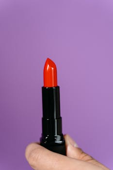 Close-up of a hand holding red lipstick against a purple background for beauty advertising.