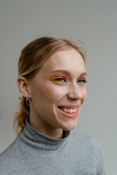 A close-up portrait of a smiling woman with vibrant eye makeup and a gray turtleneck against a neutral background.