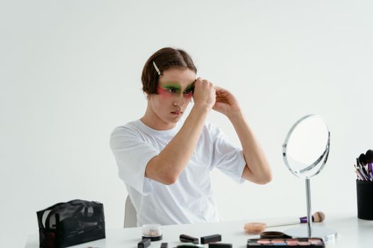 A young adult applies colorful makeup in front of a mirror indoors.