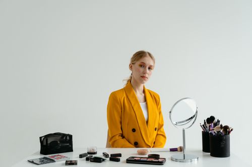 Free A woman in a yellow blazer sitting at a table with makeup tools and a mirror, ready for application. Stock Photo