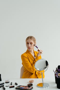 A woman in a yellow blazer applying makeup with a brush in front of a mirror indoors.
