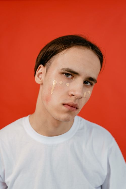 Free Young man with makeup on his face against a vibrant red background wearing a white t-shirt, looking at the camera. Stock Photo
