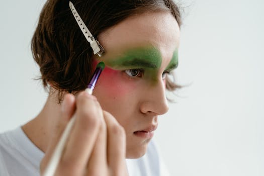Close-up portrait of a young person using makeup brush to apply vibrant eyeshadow, with a hair clip on white background.