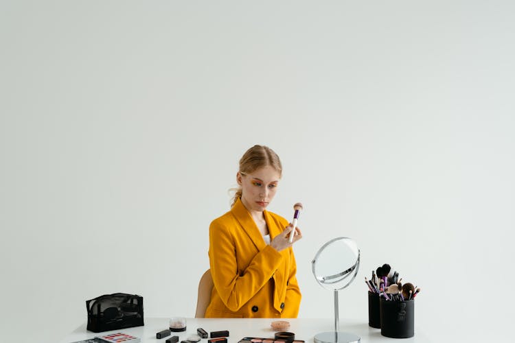 A Woman In Yellow Blazer Sitting At A Table Holding A Makeup Brush