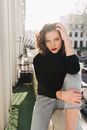 Young female in casual wear touching leg while sitting on balcony fence and looking at camera in sunny city