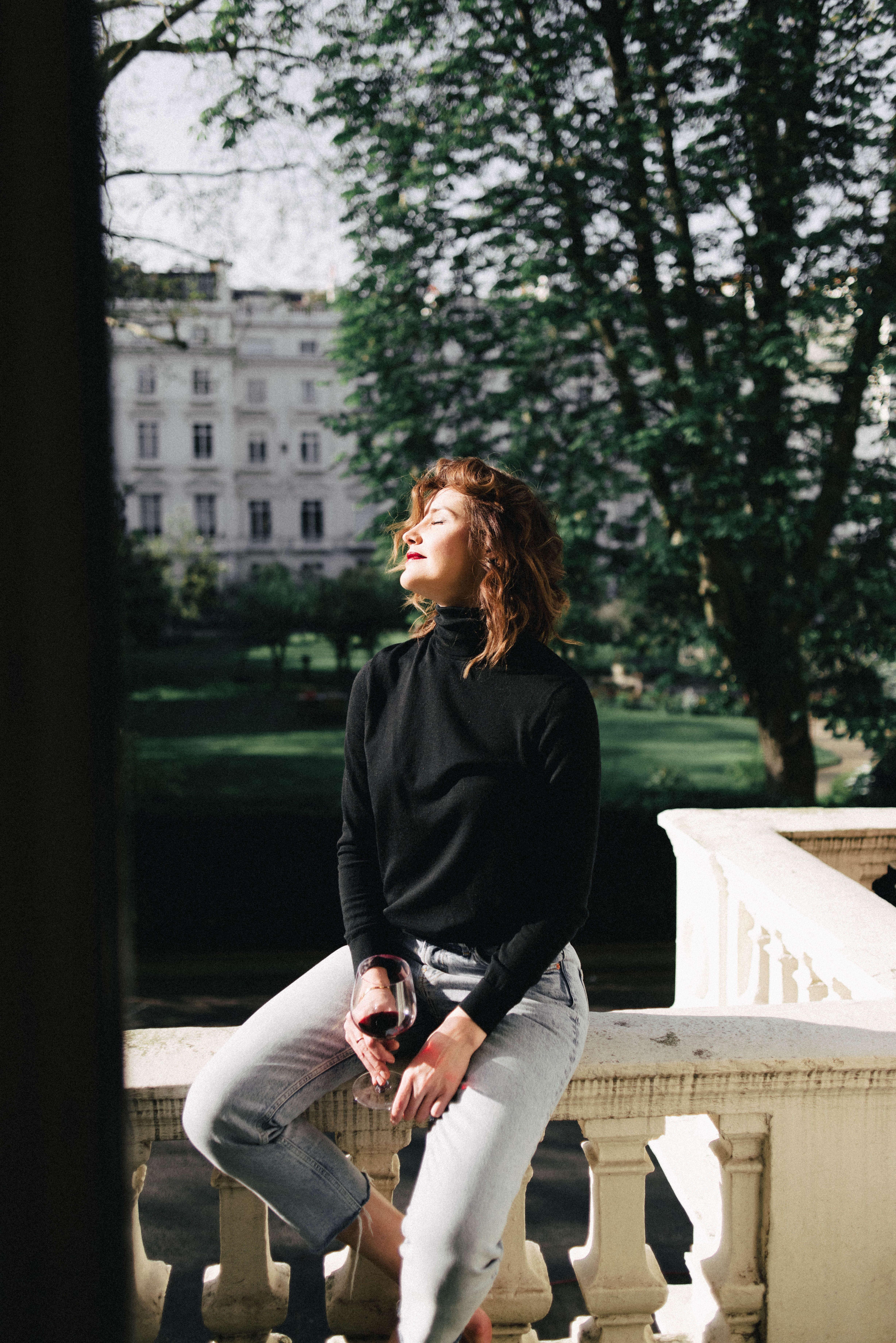 Charming young female with glass of red wine resting on balcony fence against park in sunny town
