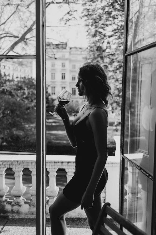 Black and white side view of young alluring female with glass of red wine looking forward at home