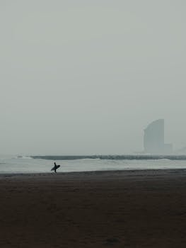 Silhouette of a surfer carrying a surfboard on a misty beach at dawn with waves in the background.