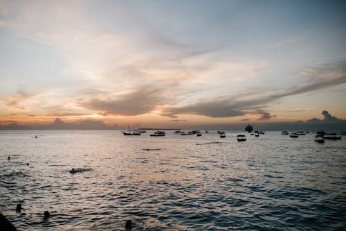 Silhouettes of faceless people swimming in ocean near boats under cloudy blue sky at sundown