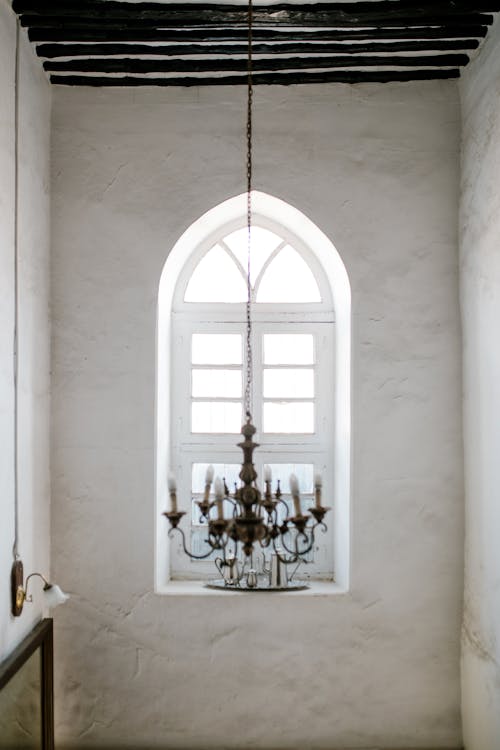 Old fashioned chandelier hanging from ceiling in room with white walls and arched window