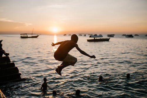 Back view of anonymous ethnic teen boy in moment of jumping in sea water at sunset