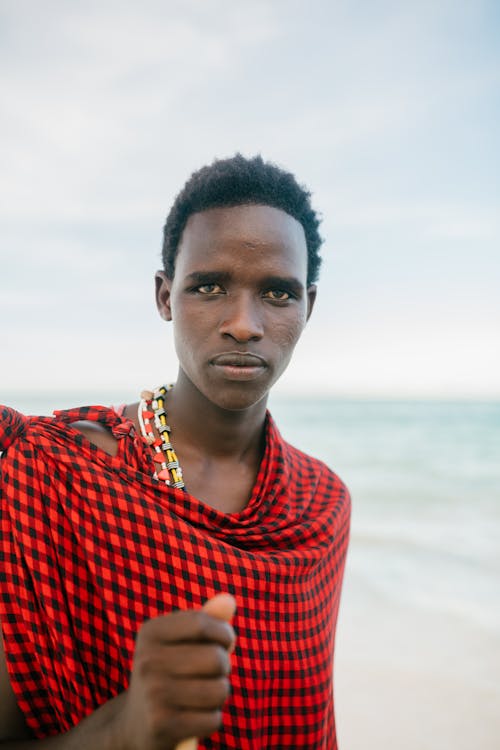 African male in traditional clothes standing on beach against sea and looking at camera