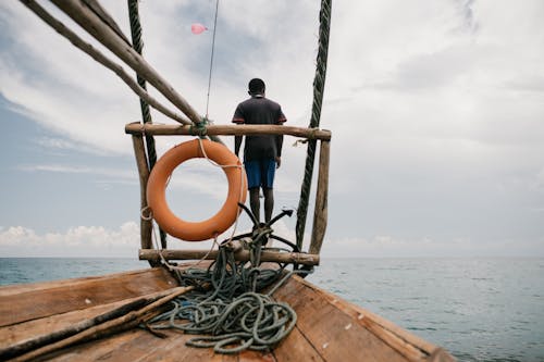 Low angle back view of ethnic male sailing shabby wooden boat in sea on cloudy day in summer