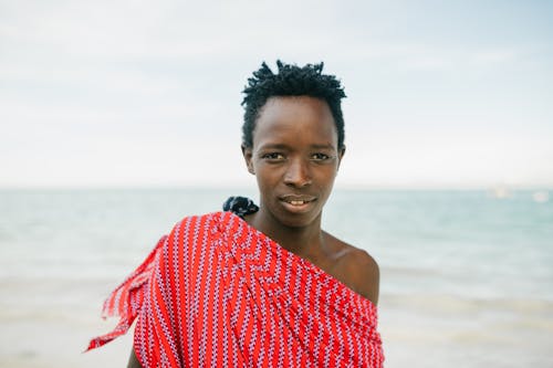Teenage African boy in red garment standing on background of sea and looking at camera