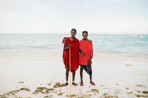 Free African father and teenage son in traditional red clothes standing on sandy beach and looking at camera Stock Photo