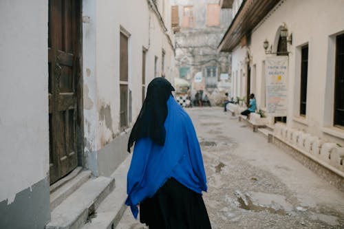 Back view of anonymous Muslim female wearing traditional hijab and cloak walking along narrow street in old city