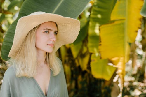 Serene female in straw hat and summer dress standing in exotic garden with banana leaves and looking away during vacation