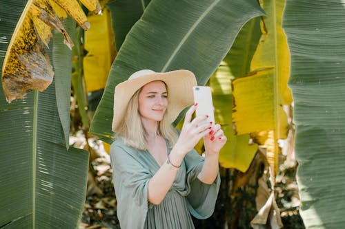 Cheerful female in straw hat standing in tropical garden and taking picture of banana leaves on smartphone while enjoying summer vacation