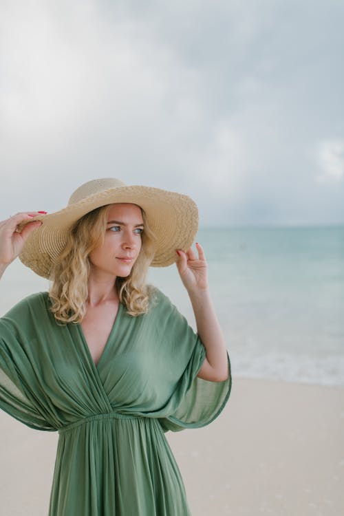 Serene female in summer dress and straw hat standing on seashore and looking away during holiday