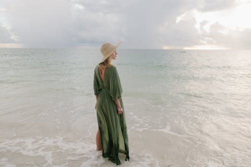 Back view of graceful female in maxi dress and straw hat standing in sea and looking away