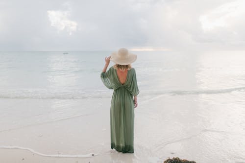 Back view of unrecognizable tender female wearing summer dress and straw hat standing on seashore and enjoying seascape during vacation