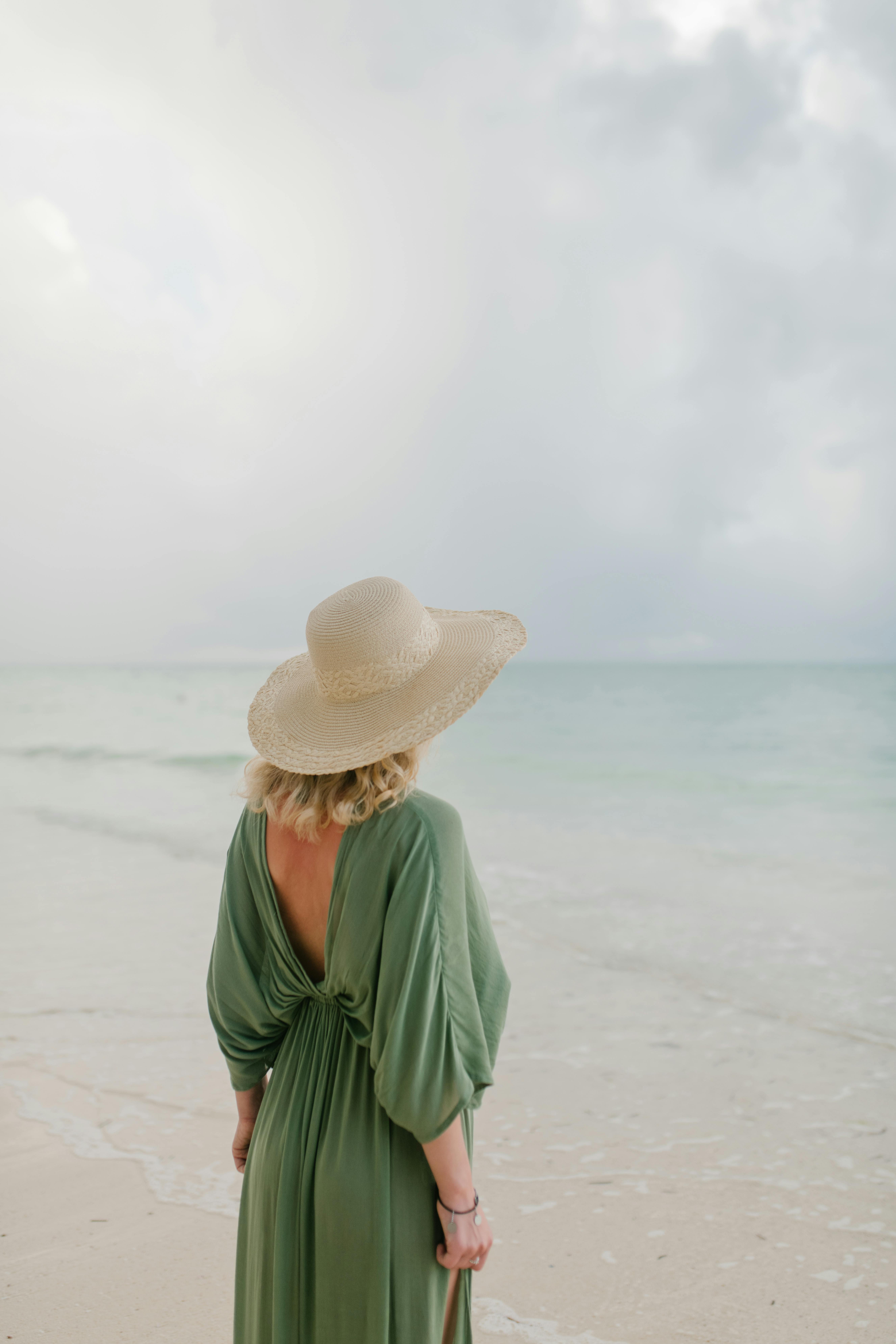 Back view of anonymous female traveler in straw hat and dress standing on sandy beach and admiring transparent sea