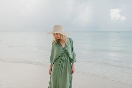Young female tourist in straw hat and dress standing on seashore washed by clear waves of sea