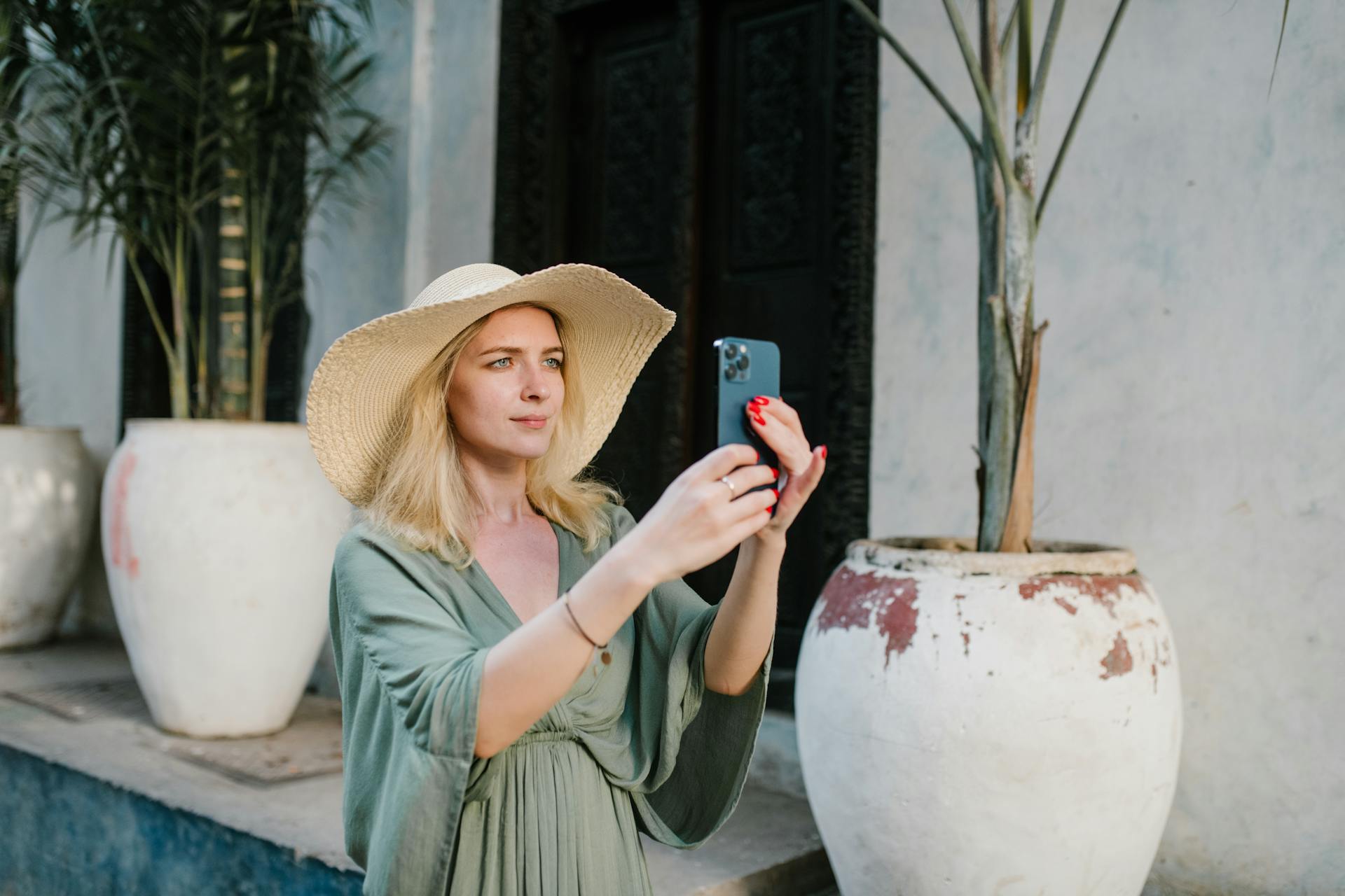 Female tourist wearing summer dress and broad brim hat taking photo on cellphone white standing on street near palms growing in clay jugs