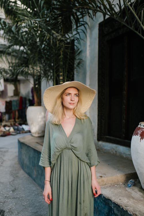 Peaceful female wearing summer dress and broad brim hat standing near palm on street of rural settlement and looking away