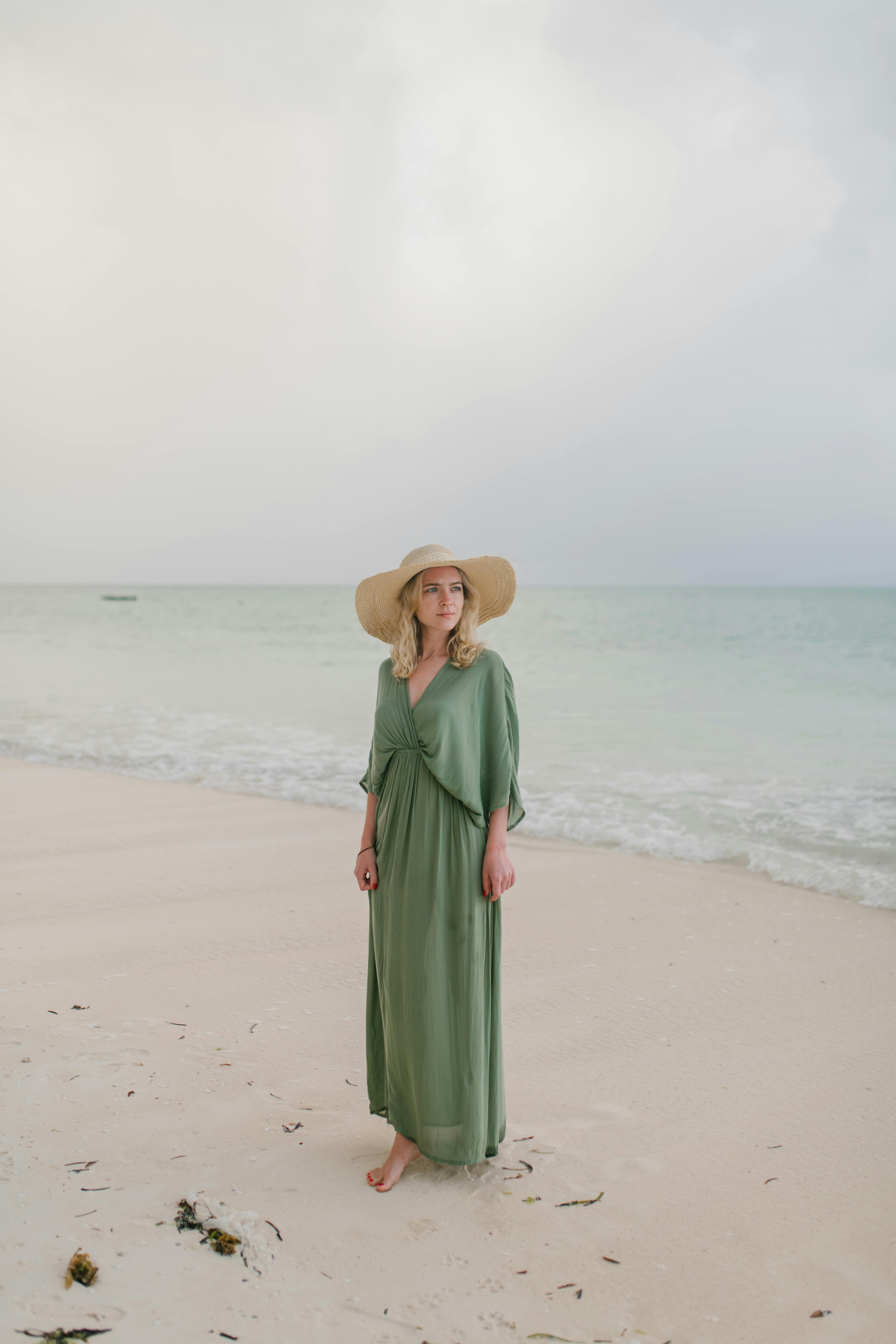 Full body of barefoot female tourist wearing long dress and straw hat standing on wet coast of clear sea under clouds