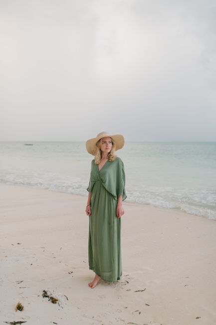 Full body of barefoot female tourist wearing long dress and straw hat standing on wet coast of clear sea under clouds