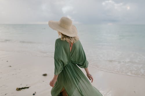 Back view of unrecognizable female tourist wearing straw hat and summer dress standing on seashore in cloudy day