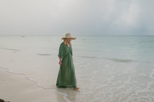 Free Full body of young female tourist wearing long summer dress and hat strolling on sandy seashore in tropical resort Stock Photo