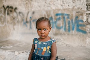 Frowning African American girl near weathered concrete building with vandal graffiti and broken wall in poor district