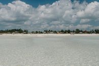 Clean transparent seawater washing sandy coastline with green plants and wooden bungalow under clouds on blue sky at tropical resort