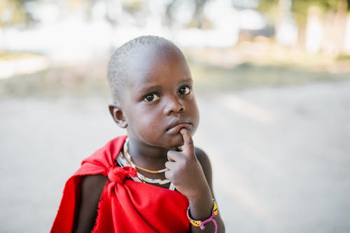 Curious black child in red cover touching chin and looking at camera with interest while standing on sunny street