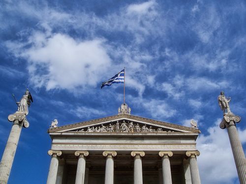 Free Front view of the Academy of Athens with Greek flag and statues against a vibrant sky. Stock Photo
