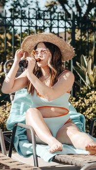 A woman in a swimsuit enjoying a summer day by the pool, sipping a drink and wearing a sun hat.